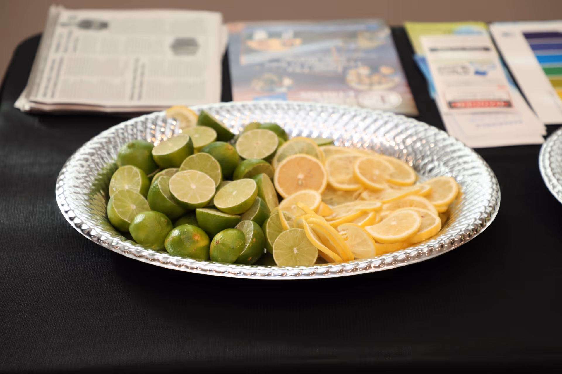 A silver platter filled with halved limes and sliced lemons placed on a black tablecloth. In the background, there are some blurred papers and brochures on the table.