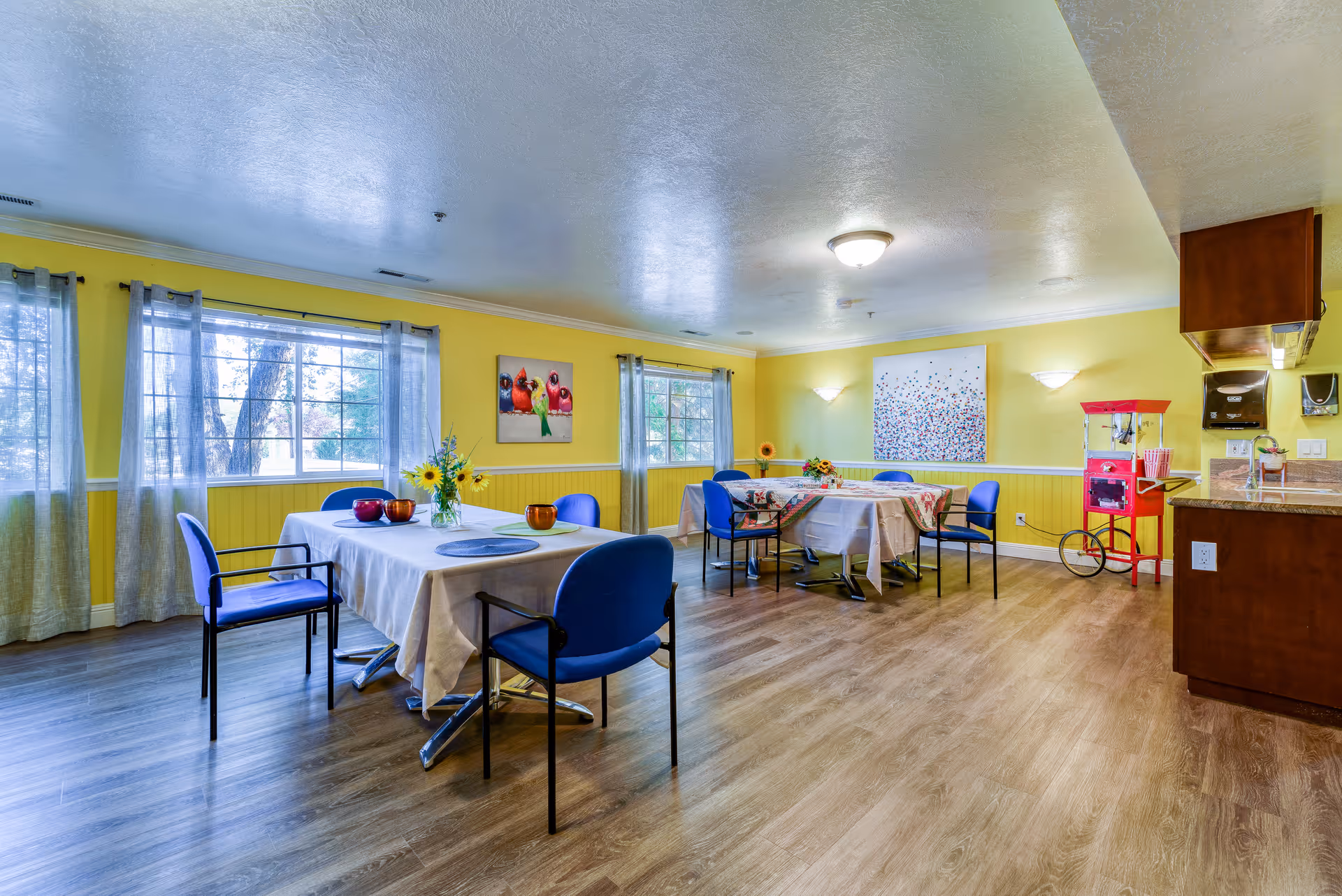 A bright dining room with yellow walls and large windows covered with sheer curtains. There are two tables covered with tablecloths, each surrounded by blue chairs. The tables have decorative items including flowers and bowls. On the right side, there is a red popcorn machine and a kitchen counter with cabinets and a sink. The floor is wooden, and colorful artwork is hung on the walls.