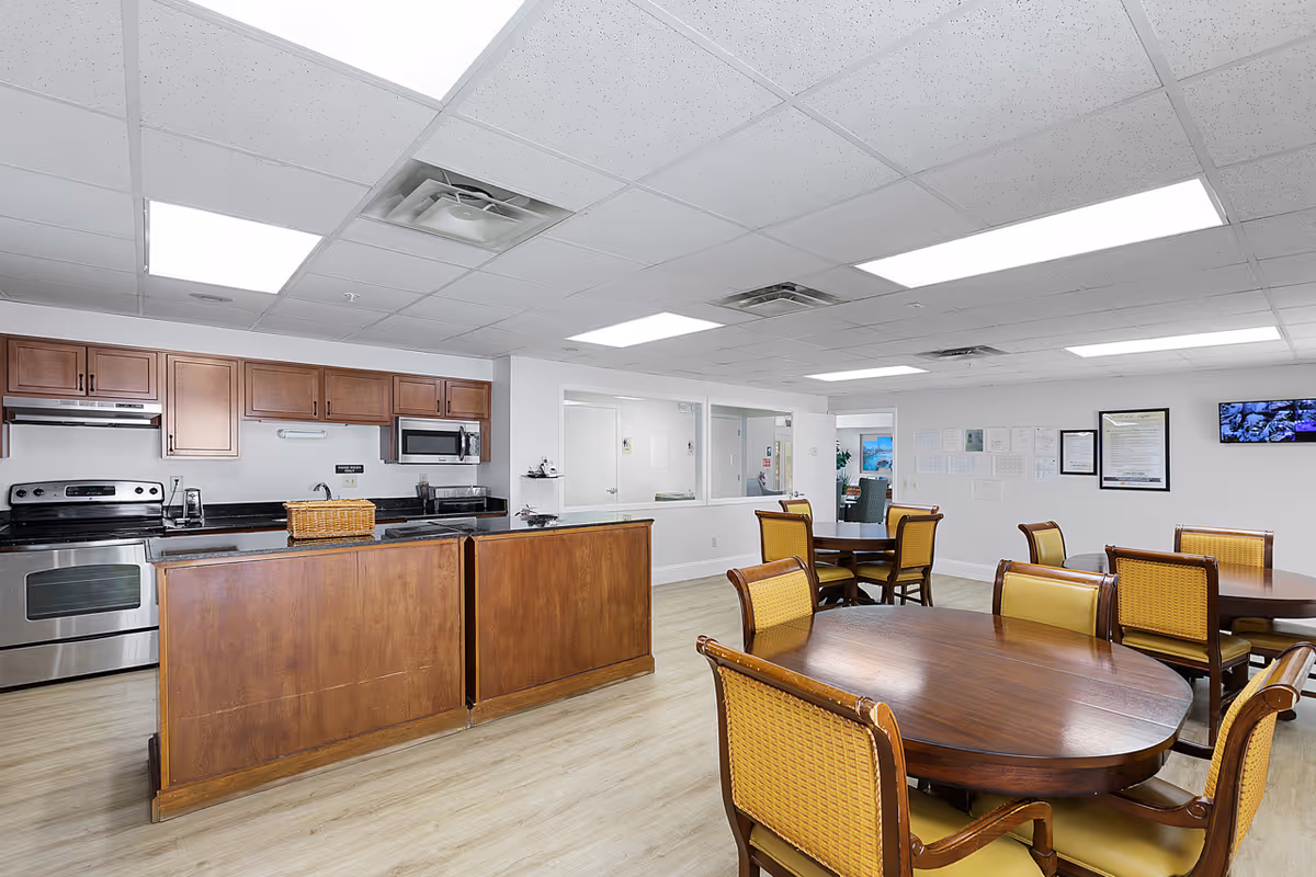 A spacious dining area with several wooden tables and chairs with yellow upholstery. In the background, there is a kitchen with wooden cabinets, stainless steel appliances including an oven and microwave, and a countertop with a basket on it. The room has a white ceiling with fluorescent lighting and light-colored wooden flooring.