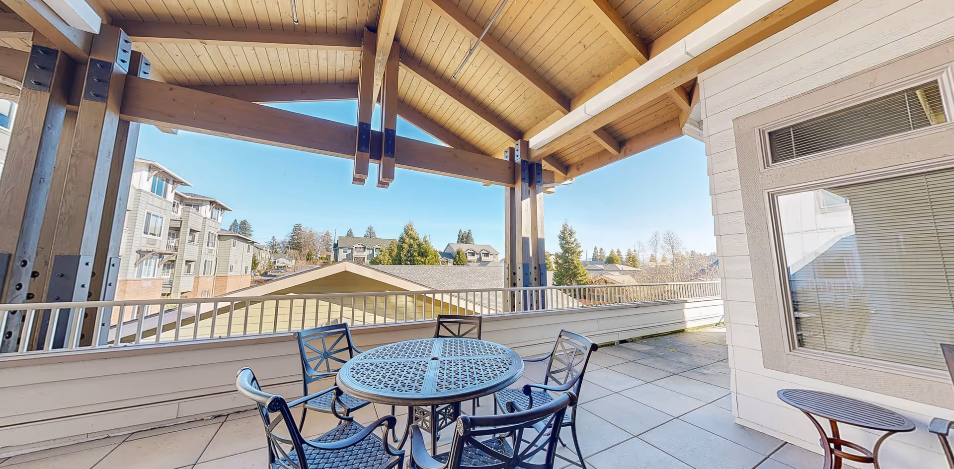 Covered outdoor patio area with a round metal table and four matching chairs. The patio has a tiled floor and wooden beams supporting the roof. In the background, there are residential buildings and trees under a clear blue sky.