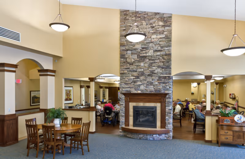 A spacious common area in a senior living facility featuring a large stone fireplace in the center. There are wooden tables and chairs arranged around the room, with several elderly residents seated and engaged in activities or conversation. The walls are painted a warm beige color, and the ceiling has hanging light fixtures. The room has a cozy and welcoming atmosphere.