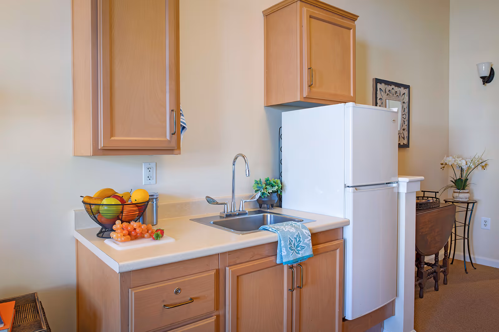 Small kitchenette with a sink and faucet, white refrigerator, wooden cabinets, and a fruit bowl on the countertop.