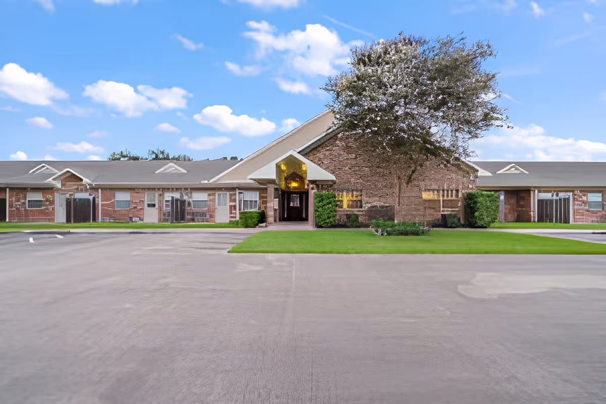Front exterior view of Morada Victoria facility showing a single-story brick building with a central entrance, green lawn, and a large tree under a blue sky with scattered clouds.