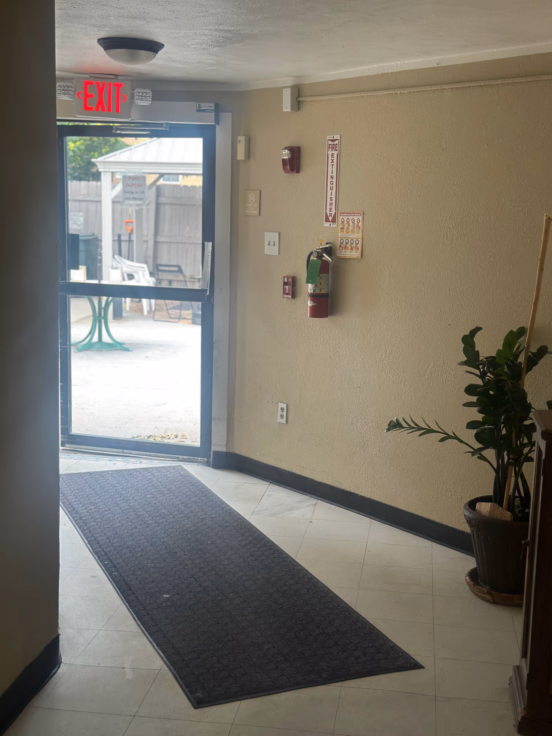 Interior entryway with an open glass exit door, a floor mat, wall-mounted fire extinguisher, and a potted plant.