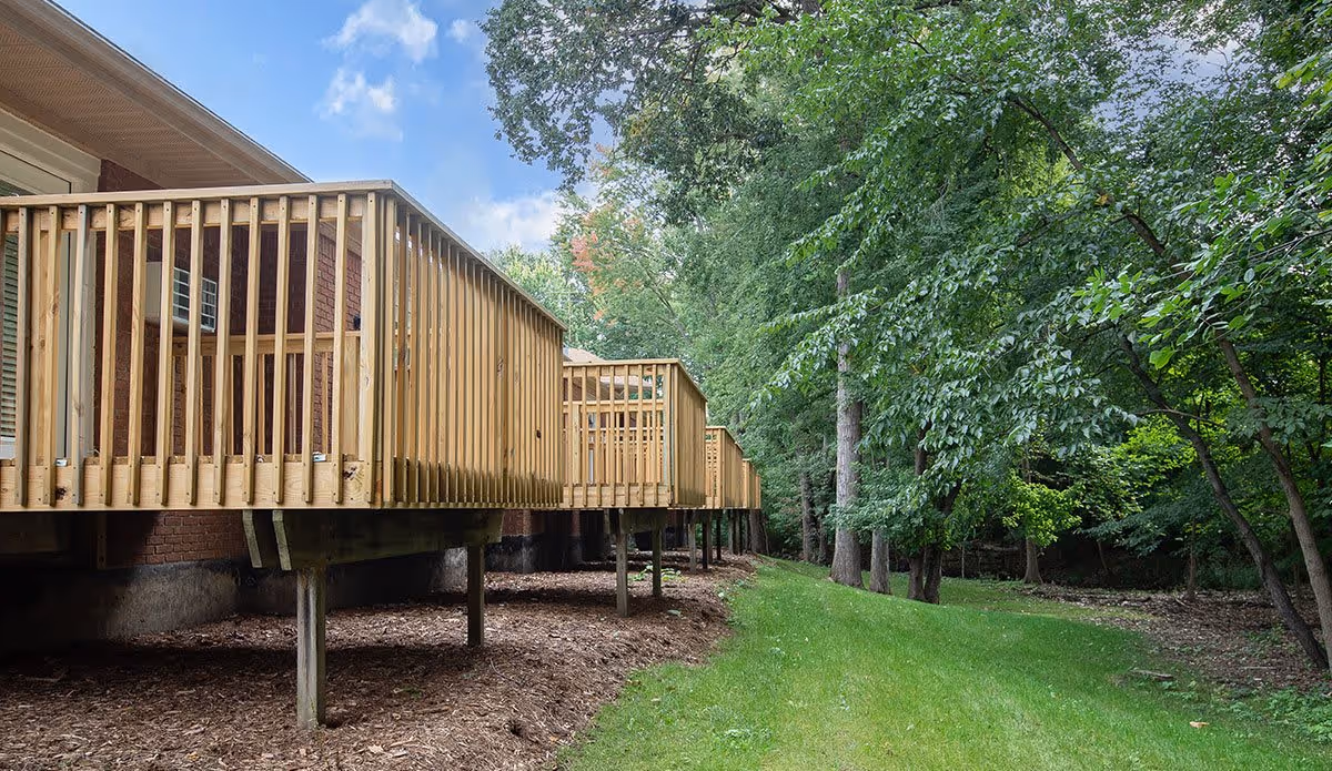View of the back side of a building with multiple wooden balconies elevated on posts, adjacent to a grassy area and surrounded by tall green trees under a partly cloudy sky.