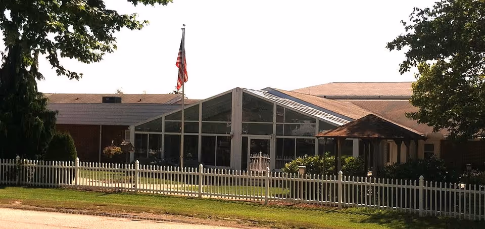 Front exterior of a single-story care facility featuring a glass atrium entrance, an American flag, a white picket fence, and a small gazebo.