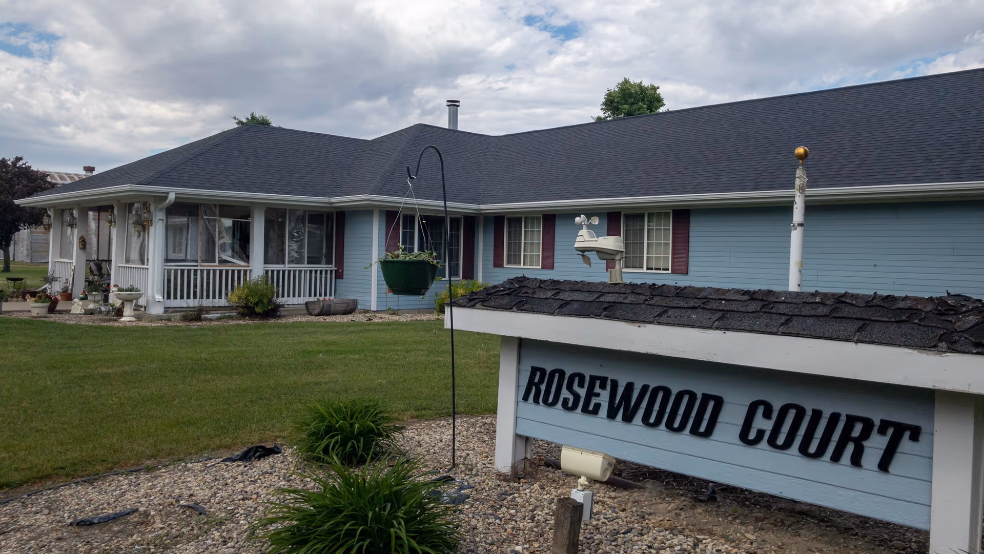 Exterior view of a single-story residential building with light blue siding and a dark shingled roof under a cloudy sky. In the foreground, there is a sign that reads 'ROSEWOOD COURT' surrounded by landscaping with grass and small plants.