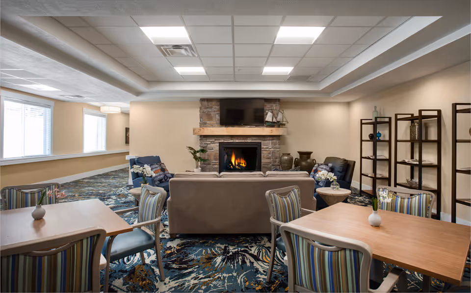 A cozy common area in a senior living facility featuring a stone fireplace with a wooden mantel and a flat-screen TV mounted above it. The room has a beige sofa and two blue armchairs arranged around the fireplace. There are two wooden tables with striped cushioned chairs in the foreground, each table decorated with a small vase of flowers. The room has patterned carpet, beige walls, and ceiling lights. Shelving units with decorative items are visible on the right side.
