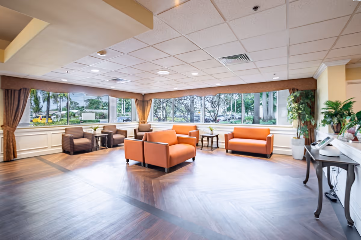 A spacious and well-lit common area with large windows showing outdoor greenery. The room features orange cushioned sofas and armchairs arranged around small wooden tables. The floor has a wood pattern, and there are plants and a small table with a device on the right side.