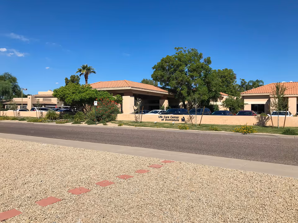 Exterior view of Life Care Center of Scottsdale building with beige walls and terracotta roof tiles, surrounded by trees and parked cars under a clear blue sky.
