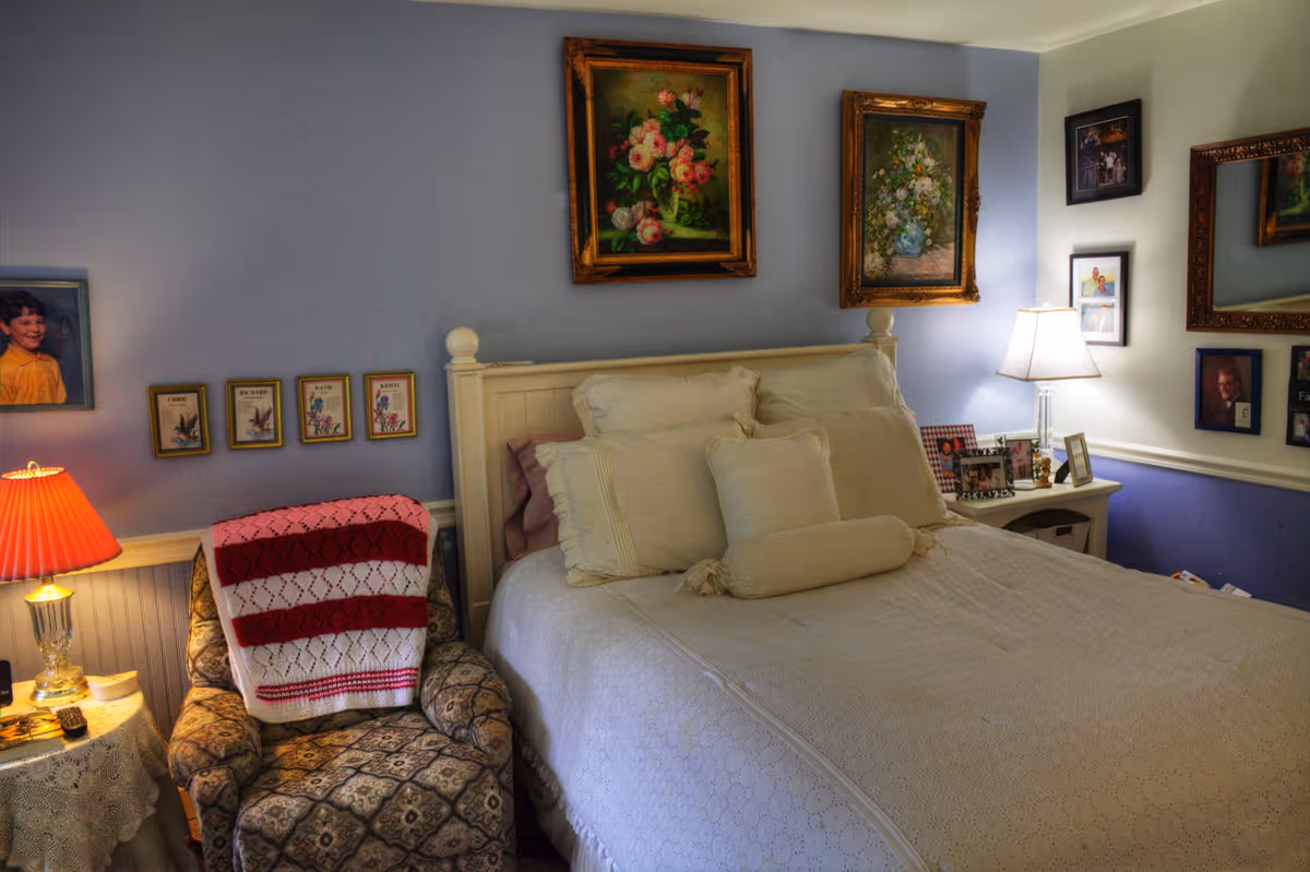 A cozy bedroom with a neatly made bed featuring multiple cream-colored pillows and a white bedspread. To the left of the bed is a patterned armchair draped with a red and white knitted blanket, next to a small round table with a lace tablecloth and a lamp with a red shade. The walls are painted light blue and decorated with framed floral paintings and family photos. A bedside table with a lamp and more framed photos is on the right side of the bed.