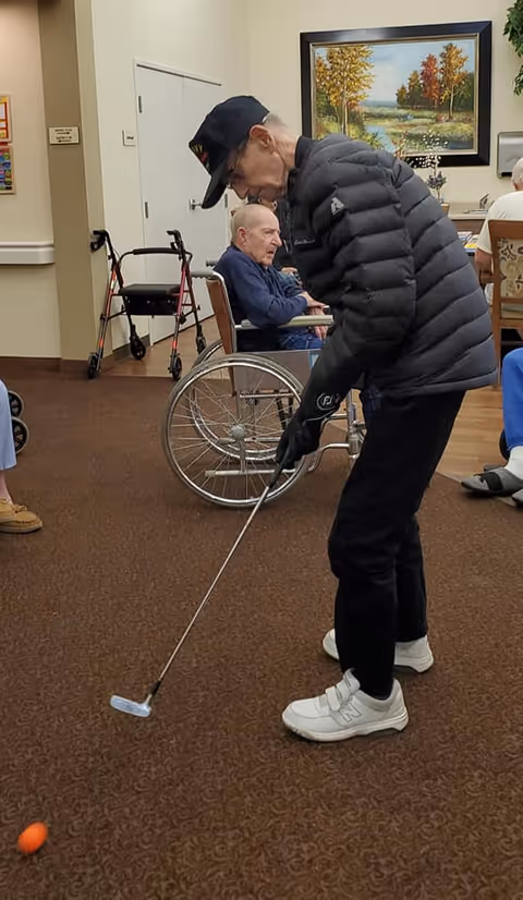 An elderly man wearing a black puffer jacket, black pants, white sneakers, and a black cap is playing indoor golf with a putter and an orange ball on a carpeted floor. In the background, another elderly man is seated in a wheelchair, and there is a walker nearby. The room has beige walls, a framed landscape painting, and other seated individuals partially visible.