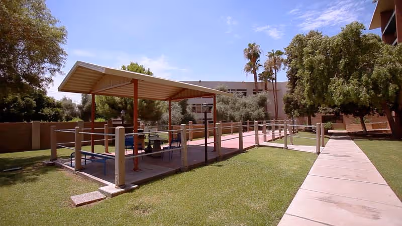 Outdoor covered seating area with tables and chairs surrounded by a grassy lawn, trees, and a paved walkway under a clear blue sky.