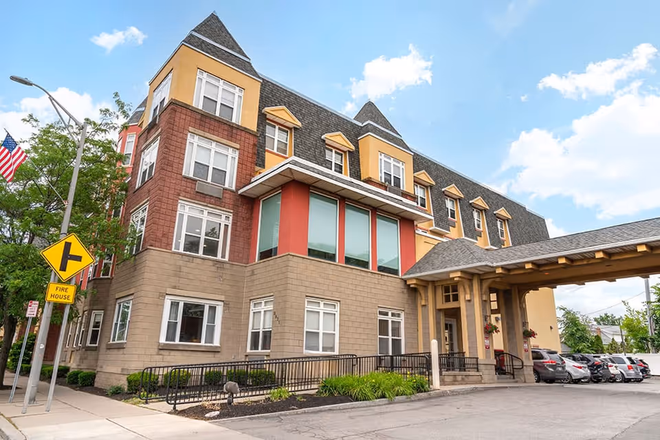 Front exterior of a multi-story senior living building with a covered porte-cochere, parked cars, and an American flag.
