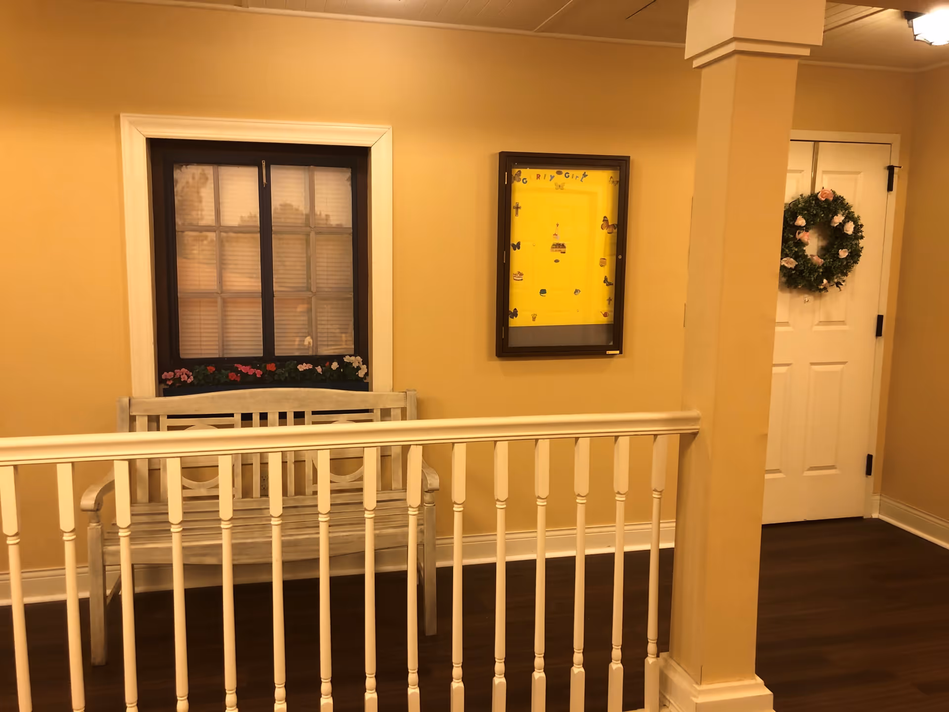 Indoor hallway area with a white wooden bench beneath a window with closed blinds and flower decorations on the windowsill. A yellow bulletin board with various small decorations is mounted on the wall next to a white door adorned with a green wreath with pink flowers. The floor is dark wood, and there is a white railing in the foreground.