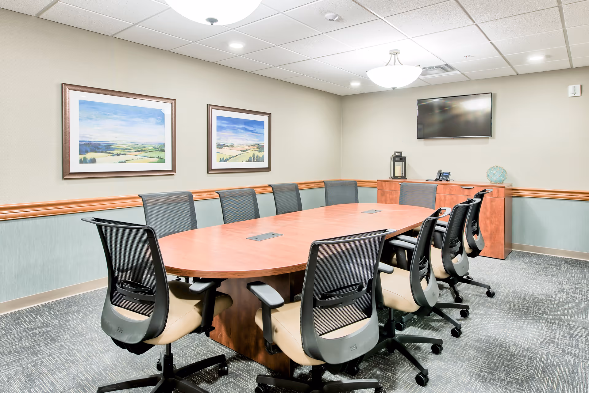 A conference room with a large oval wooden table surrounded by nine black mesh office chairs. The room has light-colored walls with a wooden chair rail, two landscape paintings on one wall, a flat-screen TV mounted on another wall, and a wooden cabinet beneath the TV holding a lantern, a phone, and a decorative glass object. The ceiling has recessed lighting and a central hanging light fixture.