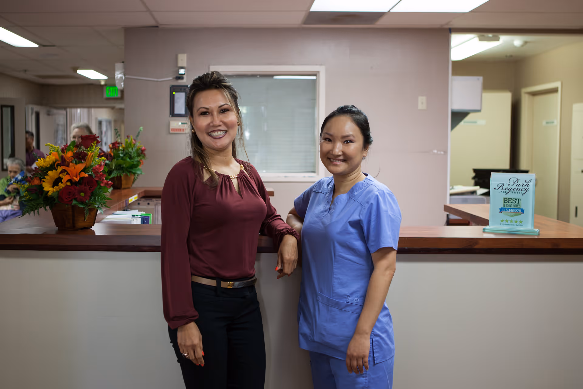 Two women standing and smiling in a care center reception area. One woman is wearing a maroon blouse and black pants, and the other is wearing blue medical scrubs. Behind them is a counter with flower arrangements and a sign that reads 'Park Regency Care Center Best Nursing Home US News & World Report'.