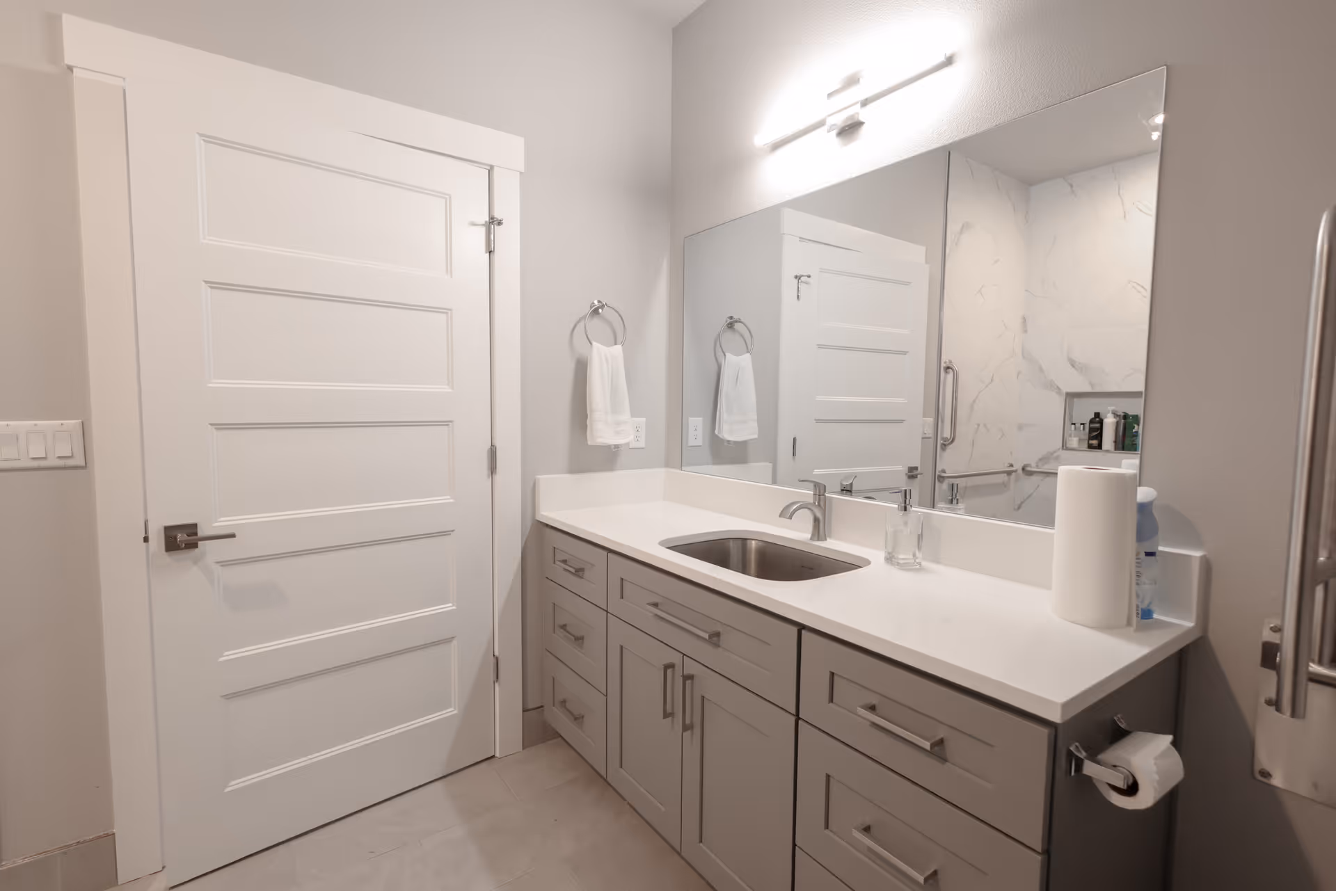 Well-lit modern bathroom with a sink vanity, large mirror, cabinets, and a door.