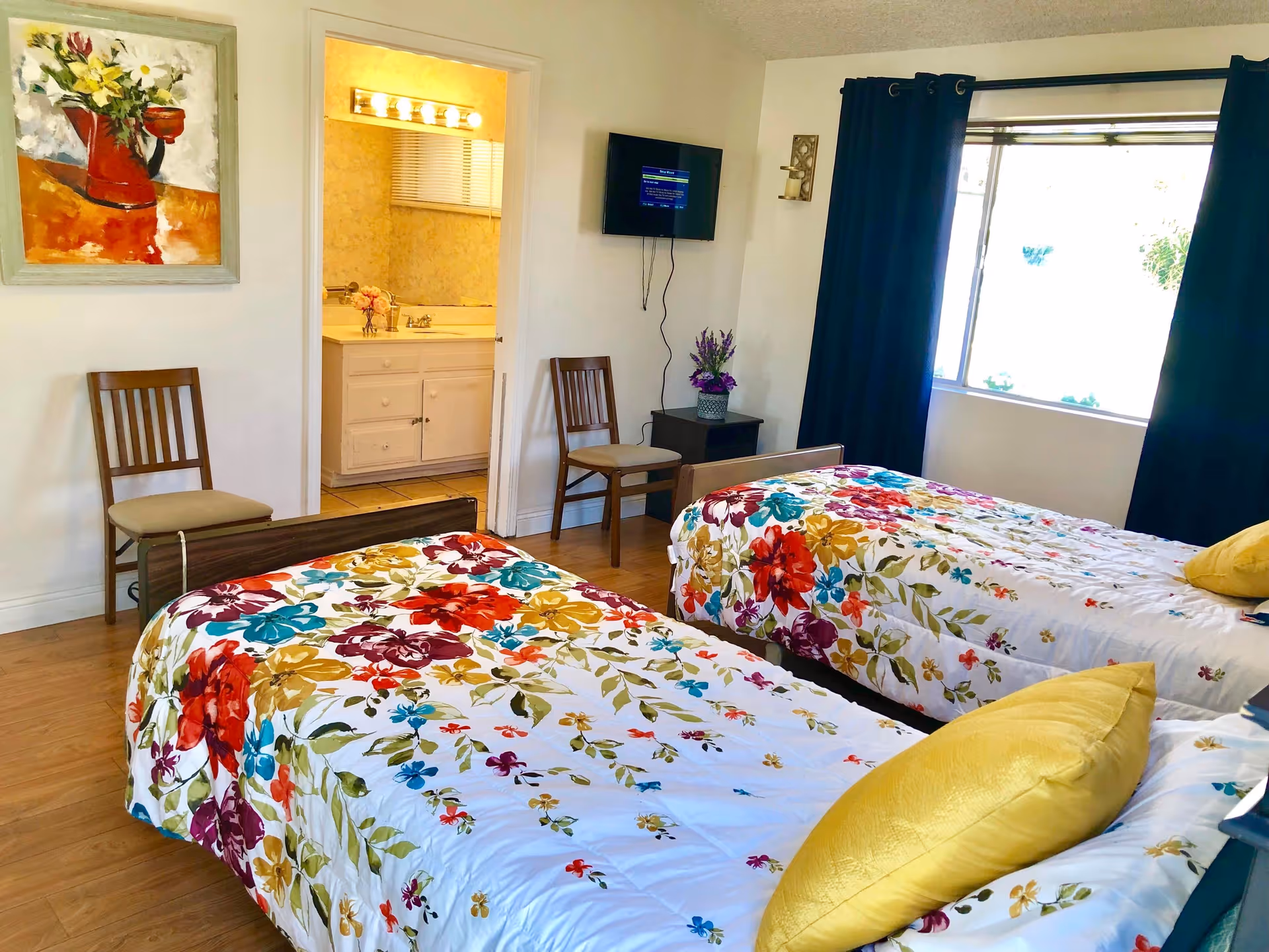 A bright bedroom with two twin beds covered in colorful floral bedspreads and yellow pillows. There are two wooden chairs, a small side table with a purple flower arrangement, a wall-mounted TV, and a large window with dark curtains. A doorway leads to a bathroom with a sink and mirror. A painting of flowers in a red vase hangs on the wall.