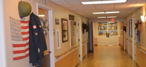 A well-lit hallway in a senior living facility with wooden flooring and beige walls. On the left wall, there is a hanging American flag, a military helmet, and a jacket with patches. The hallway has handrails on both sides and several doors leading to rooms. At the far end, there is a set of double doors with a colorful mural above them.