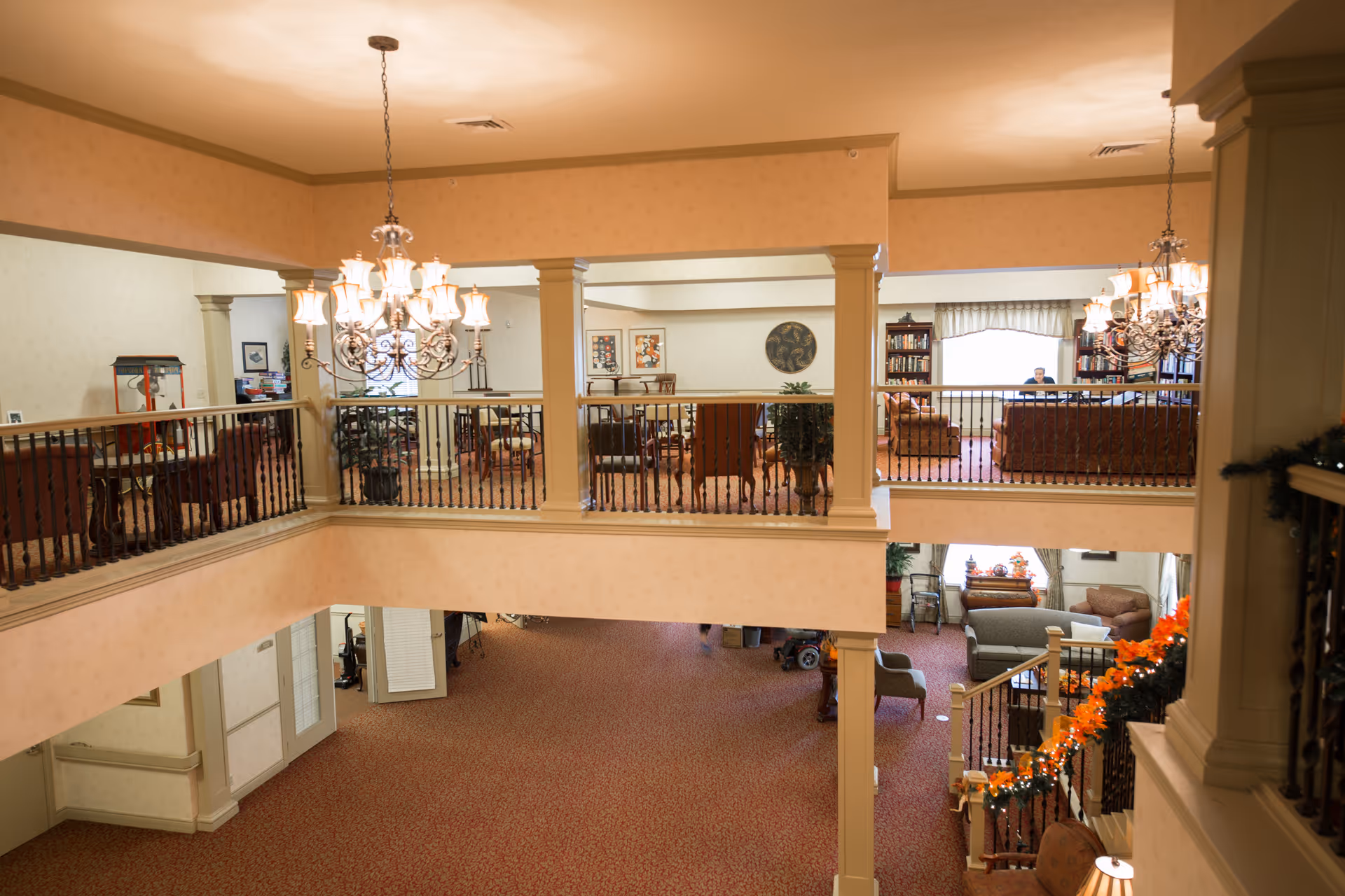 Two-story interior common area with chandeliers, railings, seating areas and a staircase in a senior living facility.