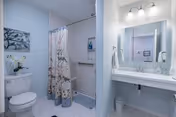 A clean and bright bathroom featuring a toilet, a shower with a floral patterned curtain, a white sink with a large mirror above it, and two wall-mounted light fixtures. The walls are painted light blue and white tiles cover the floor and shower area. A small orchid plant is placed on the toilet tank, and a towel hangs on a rack near the sink.