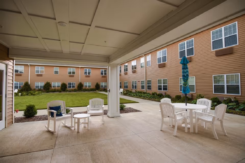 Covered patio overlooking a grassy courtyard with white chairs, a table and umbrella in front of a multi-story senior living building.