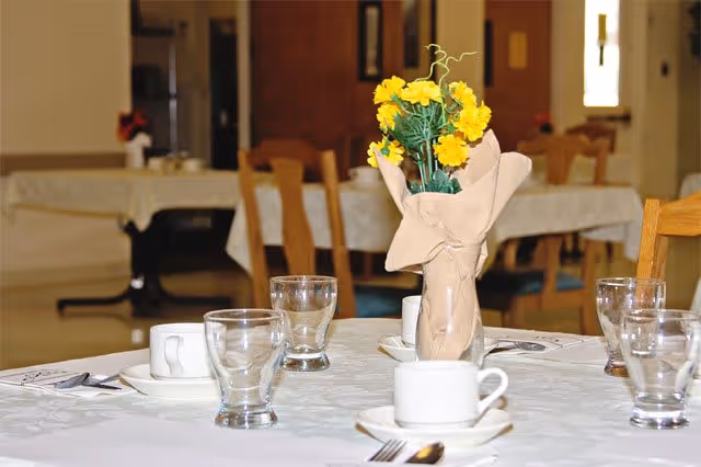 A dining table set with white tablecloth, white cups and saucers, clear drinking glasses, silverware, and a centerpiece of yellow flowers wrapped in brown paper. In the background, there are more tables and chairs in a softly lit room.