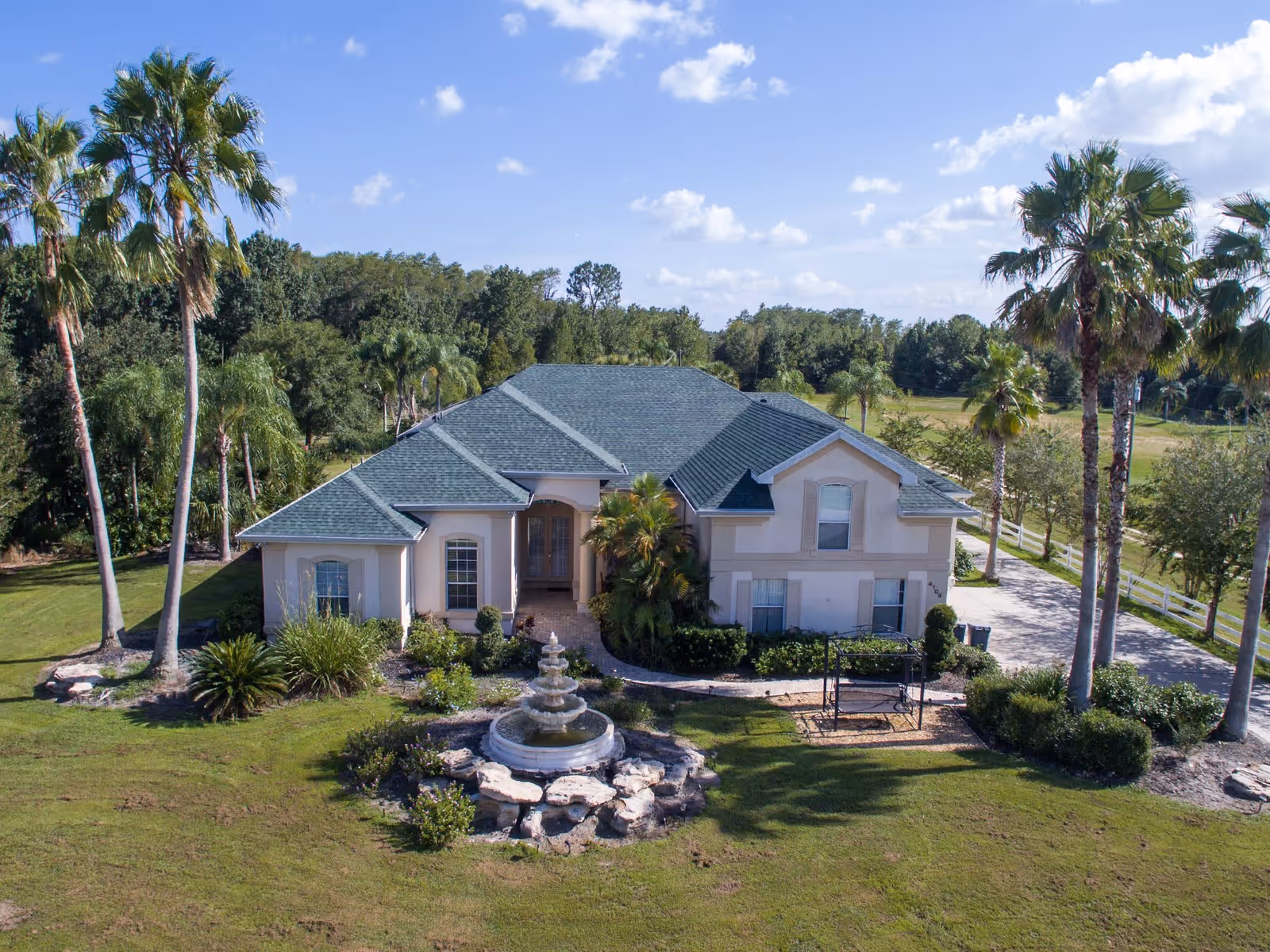 A large single-story house with a green roof surrounded by palm trees and greenery. In front of the house is a multi-tiered stone fountain with water flowing, and a paved driveway leads to the entrance. The sky is blue with scattered clouds.