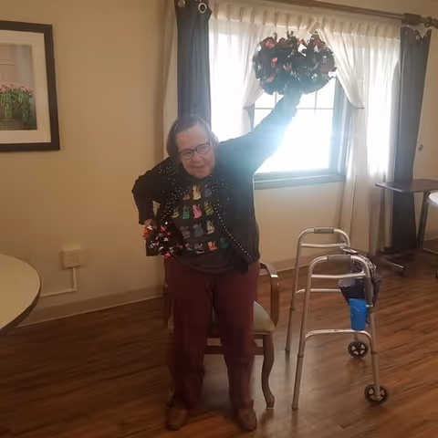 An elderly woman standing in a room with wooden flooring, holding a decorative wreath above her head. She is wearing glasses, a black cardigan with colorful cat designs, and maroon pants. Behind her is a window with white curtains, a walker with a blue cup holder, a chair, and a round table. A framed picture hangs on the wall to the left.