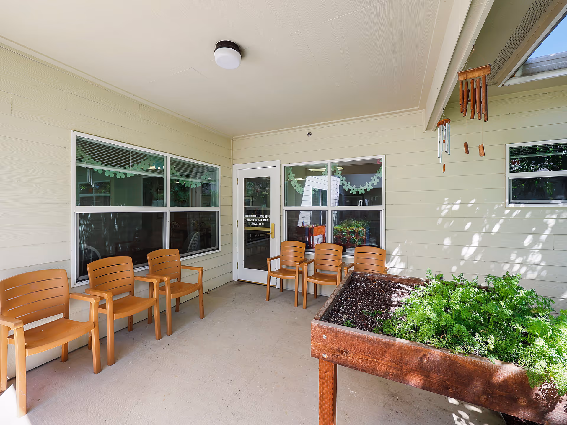 Covered outdoor patio area with six brown chairs arranged along the walls, a raised wooden planter box with green plants, and a wind chime hanging from the ceiling. The walls are light-colored with windows and a door leading inside.