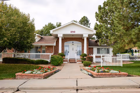 Front exterior view of a single-story brick building with white columns and a white door, surrounded by green trees and landscaped flower beds with red and white flowers. The building has the number 890 displayed above the entrance.