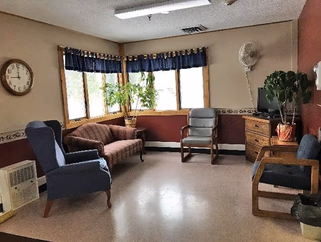 A cozy sitting area with armchairs, a small sofa, potted plants, a TV on a dresser, and bay windows.