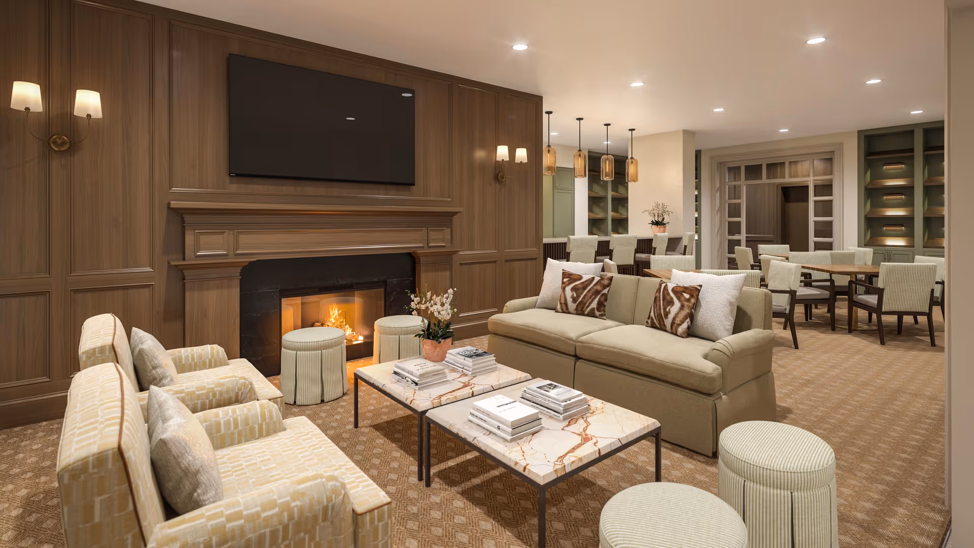 A cozy living room area in a senior living facility featuring a lit fireplace with a mounted flat-screen TV above it. The room has a beige sofa with patterned pillows, two armchairs, two marble-top coffee tables with books, and two round ottomans. In the background, there is a dining area with multiple tables and chairs, pendant lights, and built-in shelving.