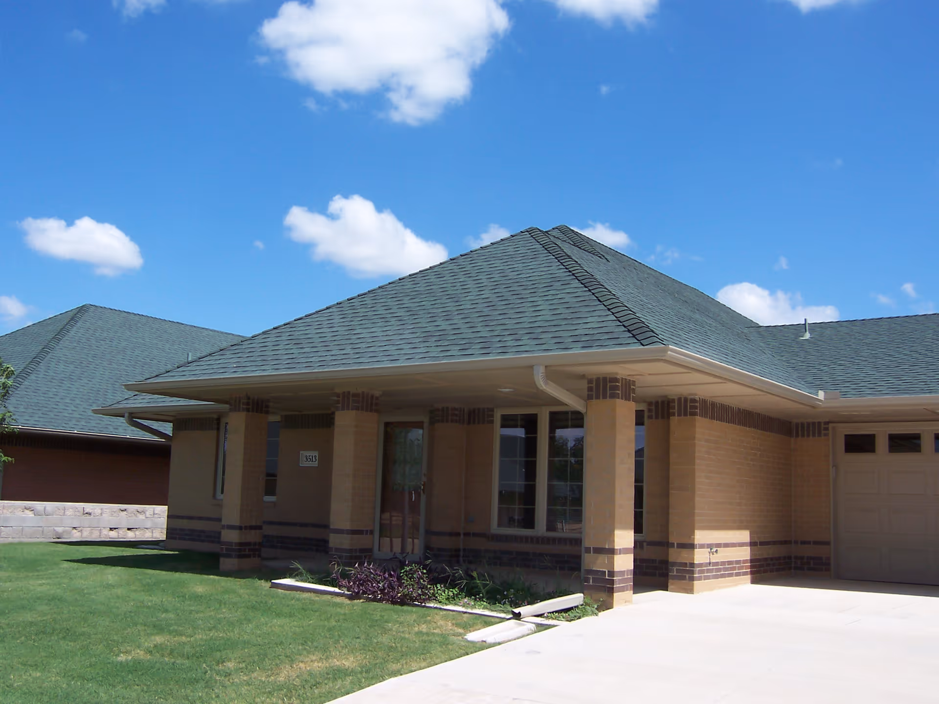 Exterior view of a single-story brick building with a green shingled roof, a covered porch supported by columns, a garage door on the right, and a well-maintained lawn under a blue sky with scattered clouds.