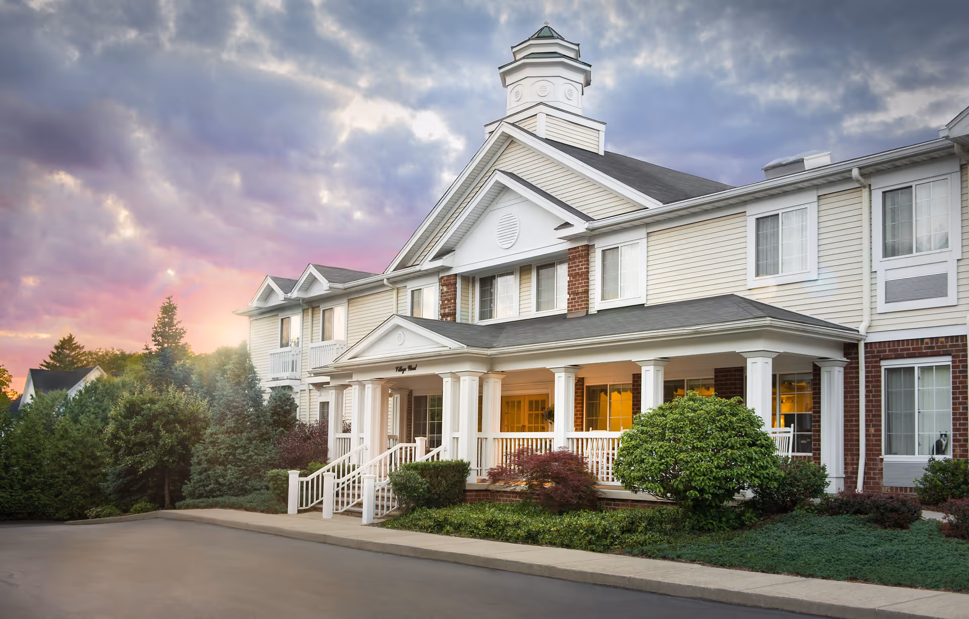 Exterior view of a senior living facility named Legacy at Village Wood during sunset, featuring a large building with white columns, beige siding, brick accents, and a cupola on the roof, surrounded by well-maintained shrubs and trees.