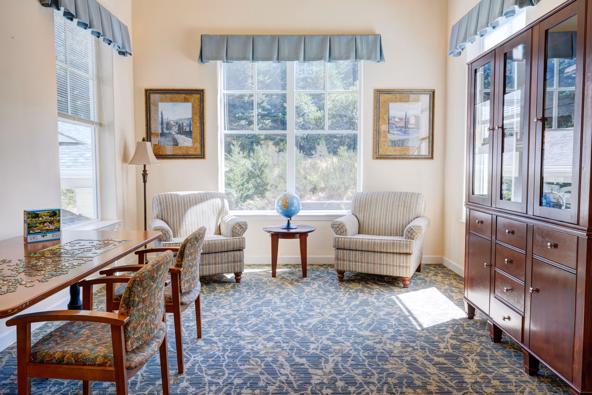 A bright sitting area with two striped armchairs facing each other with a small round wooden table holding a globe between them. The room has large windows with blue valances, framed artwork on the walls, a tall wooden cabinet with glass doors on the right, and a wooden table with two floral upholstered chairs on the left. A puzzle is spread out on the table.