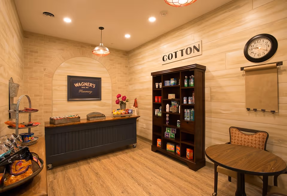 Interior view of a small pharmacy or supply room with wooden flooring and beige walls. A dark wooden counter with a sign above it reading 'WAGNER'S Pharmacy' is on the left side. On the right side, there is a tall dark wooden shelving unit labeled 'COTTON' stocked with various health and hygiene products. A round wooden table with a patterned chair is positioned near a wall clock and a paper roll dispenser.