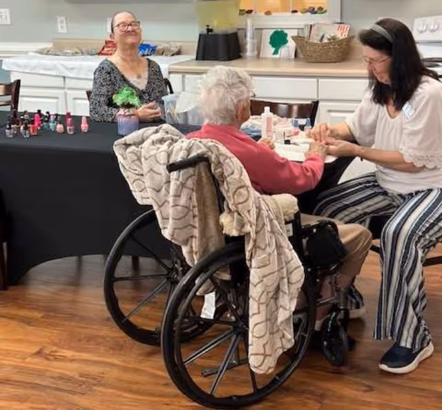 An elderly woman in a wheelchair receiving a manicure from a caregiver in a room with wooden floors. Another elderly woman sits at a table nearby with nail polish bottles and a small plant on the table. The background shows a counter with various items and a blender.
