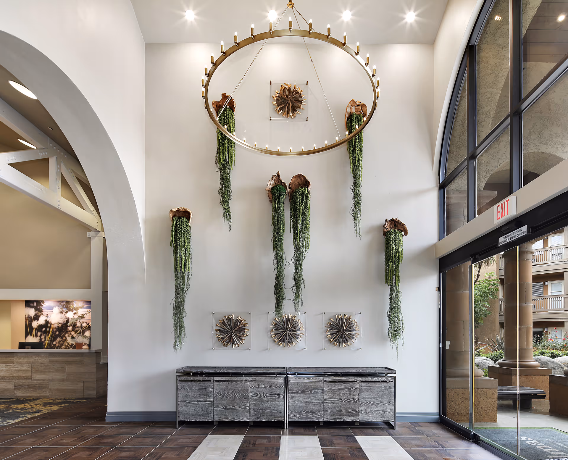 Interior view of a senior living facility lobby with a high ceiling, a large circular chandelier, and wall-mounted green plants hanging down. There is a wooden cabinet below the plants and large arched windows and doors on the right side leading outside. The floor has a patterned tile design.
