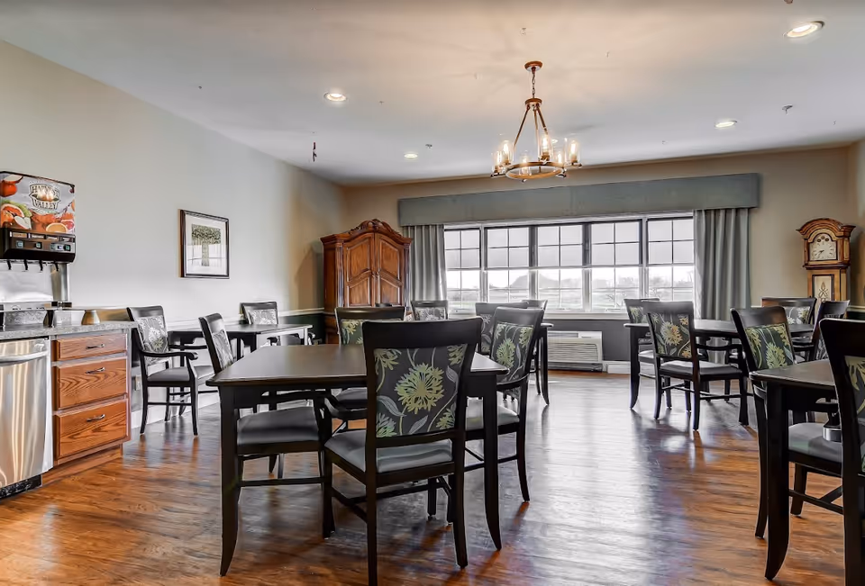 A bright dining room with several dark wooden tables and chairs featuring floral patterned backs. The room has large windows with curtains, a wooden floor, a chandelier hanging from the ceiling, a wooden cabinet, a grandfather clock, and a beverage dispenser on a countertop with drawers.