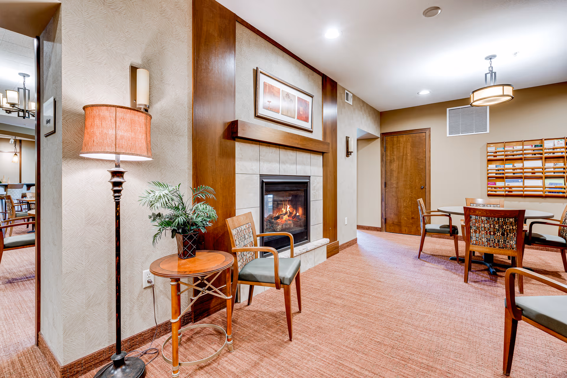A cozy interior room featuring a lit fireplace with a wooden mantel and framed artwork above it. To the left of the fireplace is a small round wooden table with a potted plant and a floor lamp with a reddish shade. Several wooden chairs with patterned cushions are arranged around the room, and a table with chairs is visible in the background. The walls are beige with a textured pattern, and the carpet is a warm reddish-brown color. A wooden door and a wall-mounted organizer with mail slots are also visible.