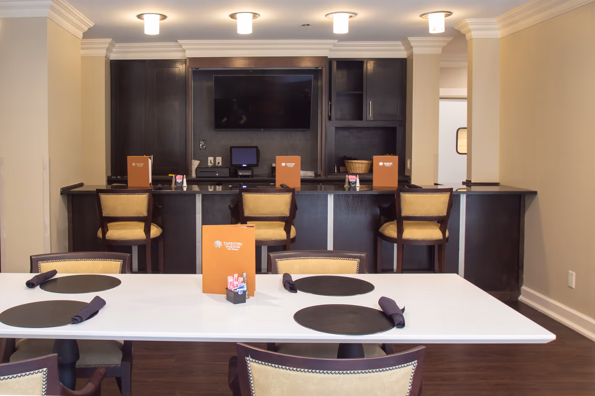 Dining area with a white table set with black placemats and napkins, surrounded by beige cushioned chairs. Behind the table is a dark wood counter with four high chairs and menus labeled 'Tapestry Senior Living'. A flat-screen TV is mounted on the wall above the counter.