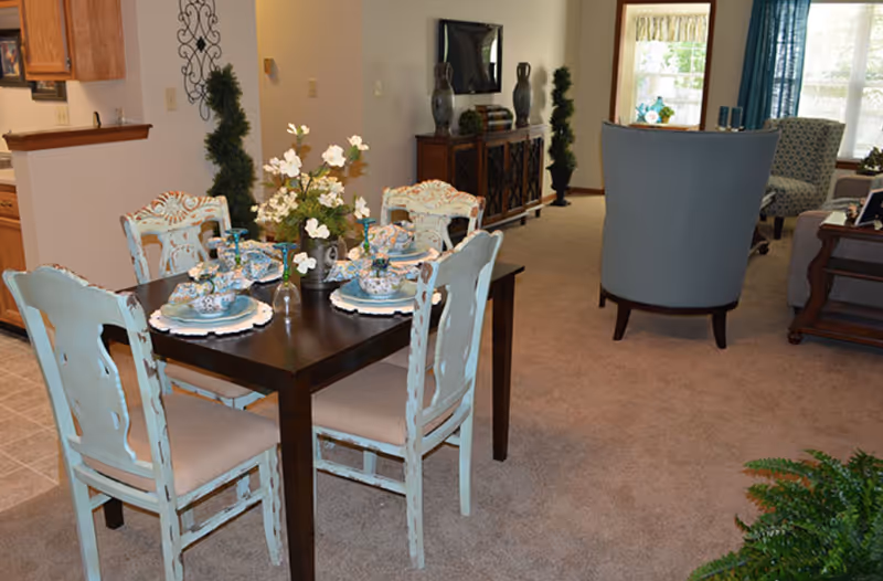 A cozy dining area with a dark wooden table set for four with floral-patterned plates, bowls, and glasses. The chairs around the table are painted in a distressed light blue finish with beige cushions. In the background, there is a living room area with upholstered chairs, a wooden sideboard with decorative items, and a window letting in natural light. The floor is carpeted, and there are some green plants and decorative topiaries around the room.