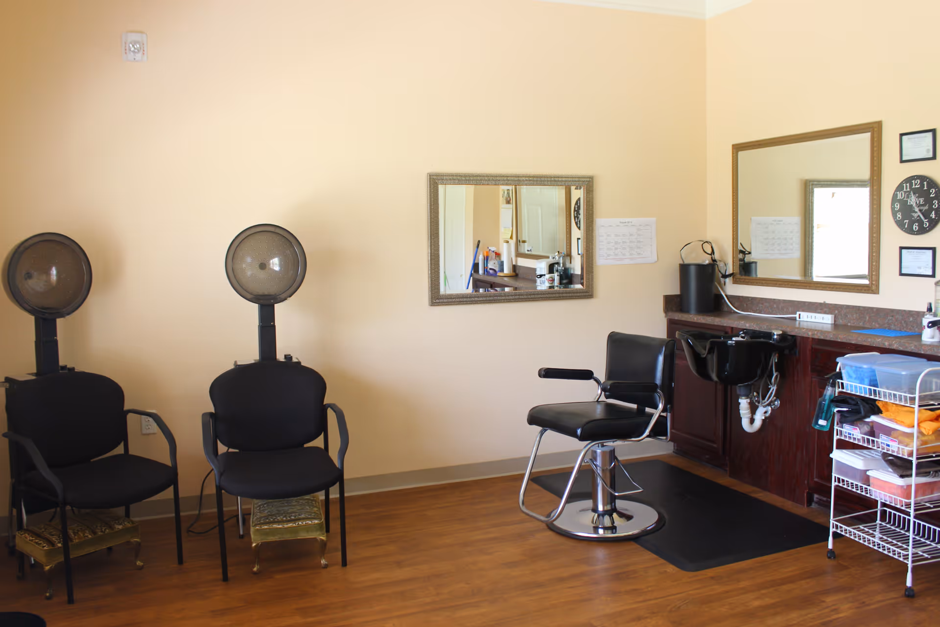 Interior of a senior living facility hair salon area with two black hair drying chairs on the left, a black salon chair in front of a sink on the right, two mirrors on the wall, and a small rolling cart with towels and supplies.