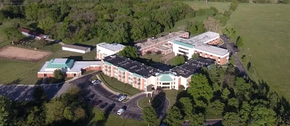 Aerial view of Our Lady Of Mercy Country Home, showing a large multi-wing building surrounded by trees and greenery, with a parking lot in front and open fields in the background.