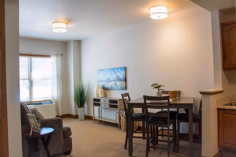 Bright furnished living and dining area with a small dining table and chairs, a sofa by a window, console table with artwork and potted plants.