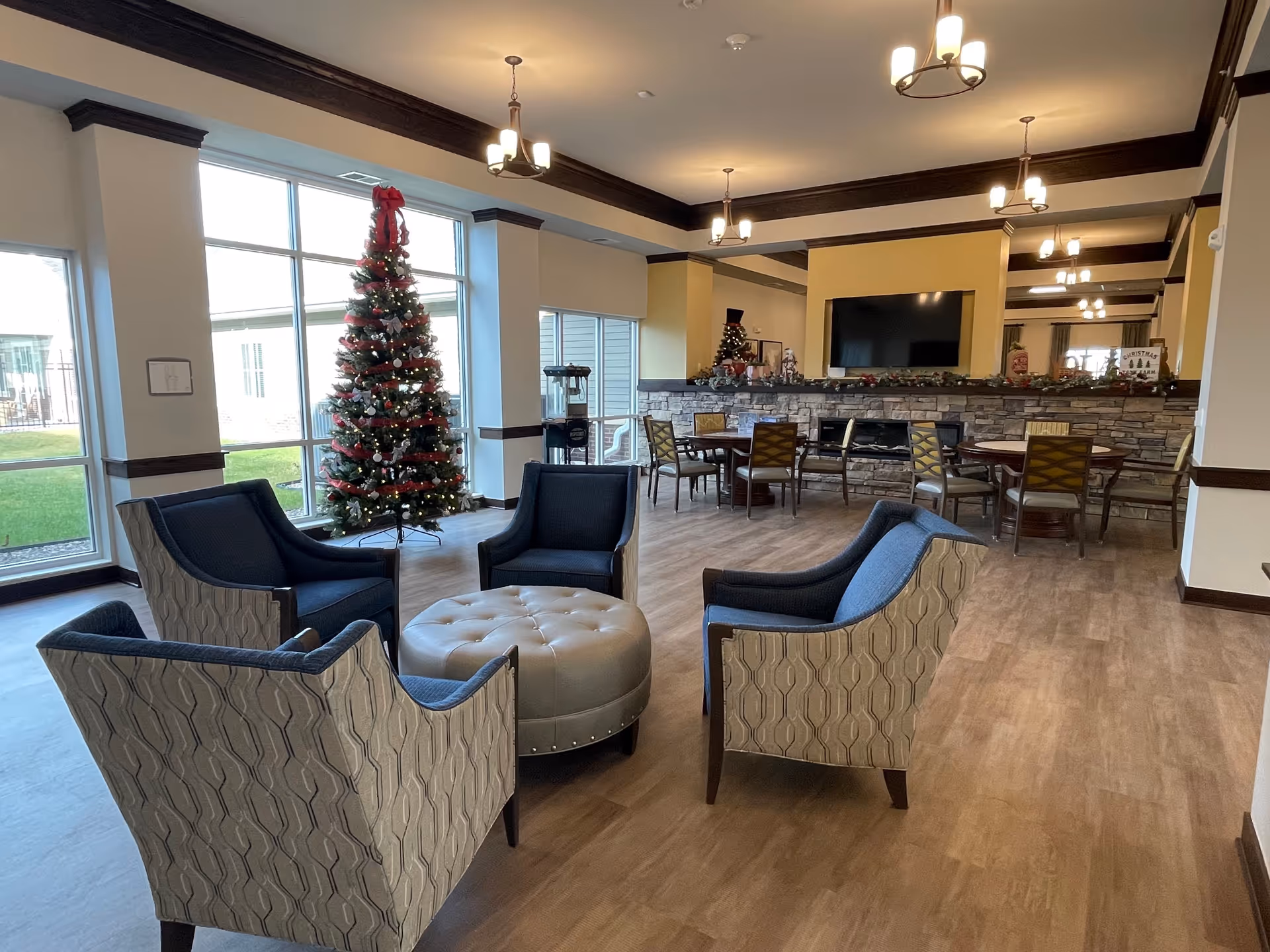 A spacious living room area decorated for Christmas with a tall Christmas tree near large windows, four upholstered armchairs arranged around a round tufted ottoman, and a stone fireplace with a mounted flat-screen TV above it. There are also several round tables with chairs in the background and festive holiday decorations on the mantel.