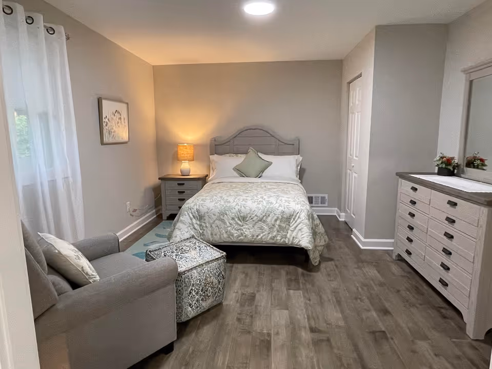 A cozy bedroom with a double bed featuring a floral bedspread and a green accent pillow. To the left of the bed is a nightstand with a lamp, and next to it is a comfortable armchair with a patterned ottoman. On the right side of the room is a dresser with a mirror and small decorative plants. The room has wooden flooring, light-colored walls, and a window with sheer white curtains.