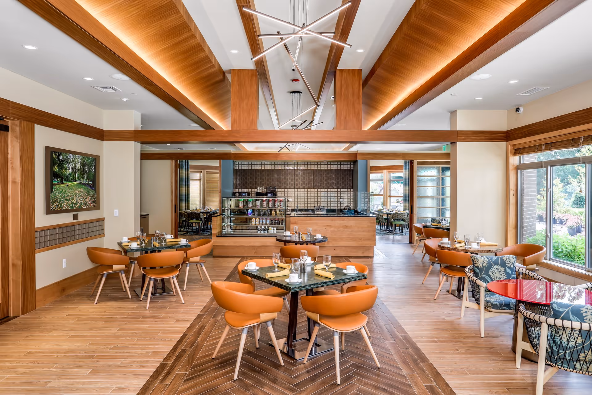 A bright and modern dining area with wooden floors and ceiling accents. The room features several tables set with glasses, cups, and napkins, surrounded by orange cushioned chairs. Large windows on the right side allow natural light to fill the space, and a display counter with food items is visible in the background. The overall design is warm and inviting with a mix of wood and neutral tones.